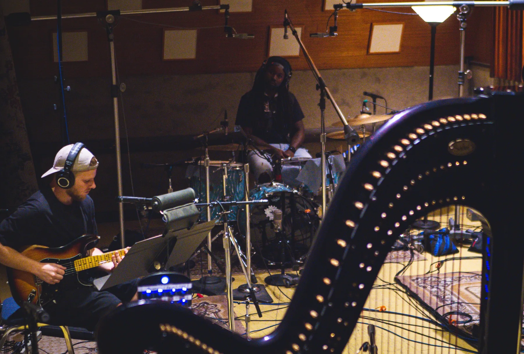 Two musicians performing in a recording room, with one playing electric bass in the foreground, another seated at drums, and a large harp crossing the right side.